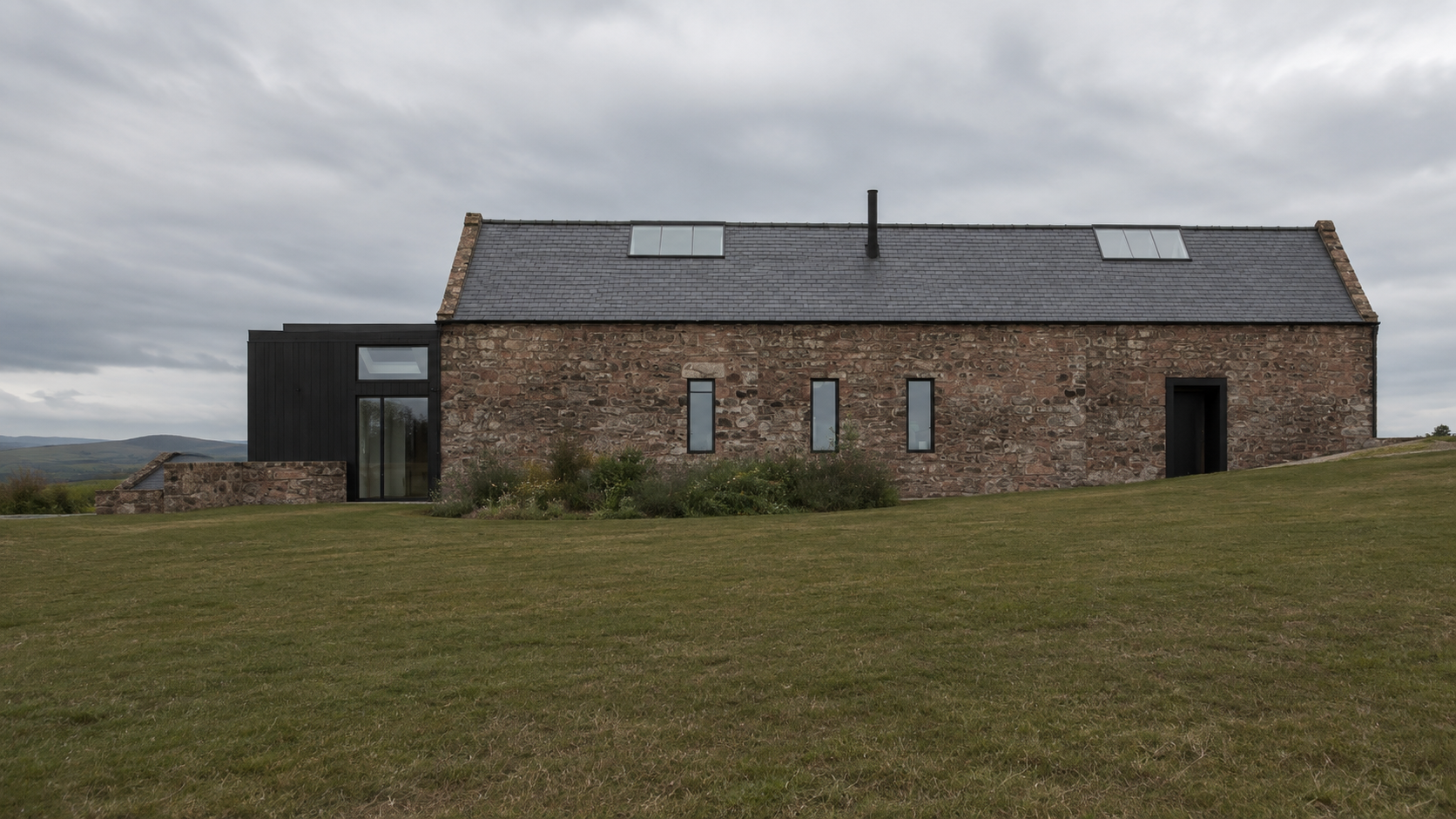 Contemporary black-clad extension meeting the original stone barn, with a glazed entrance overlooking the surrounding countryside.